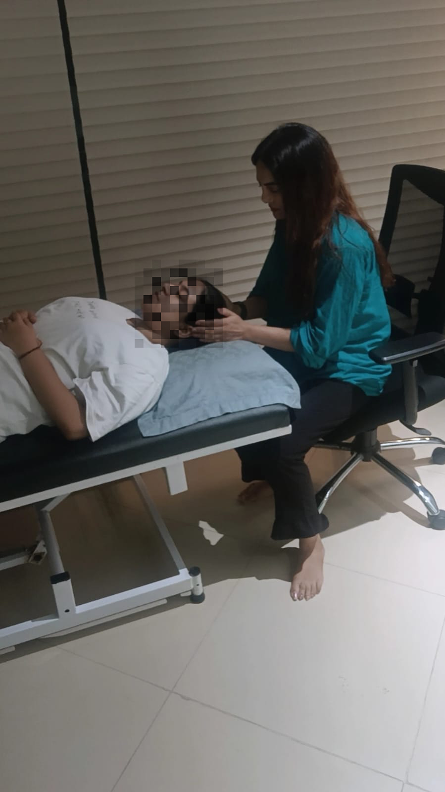 Vibha Jain seated beside a client resting on a treatment couch during a guided session. The client's face has been obscured to protect their privacy.
