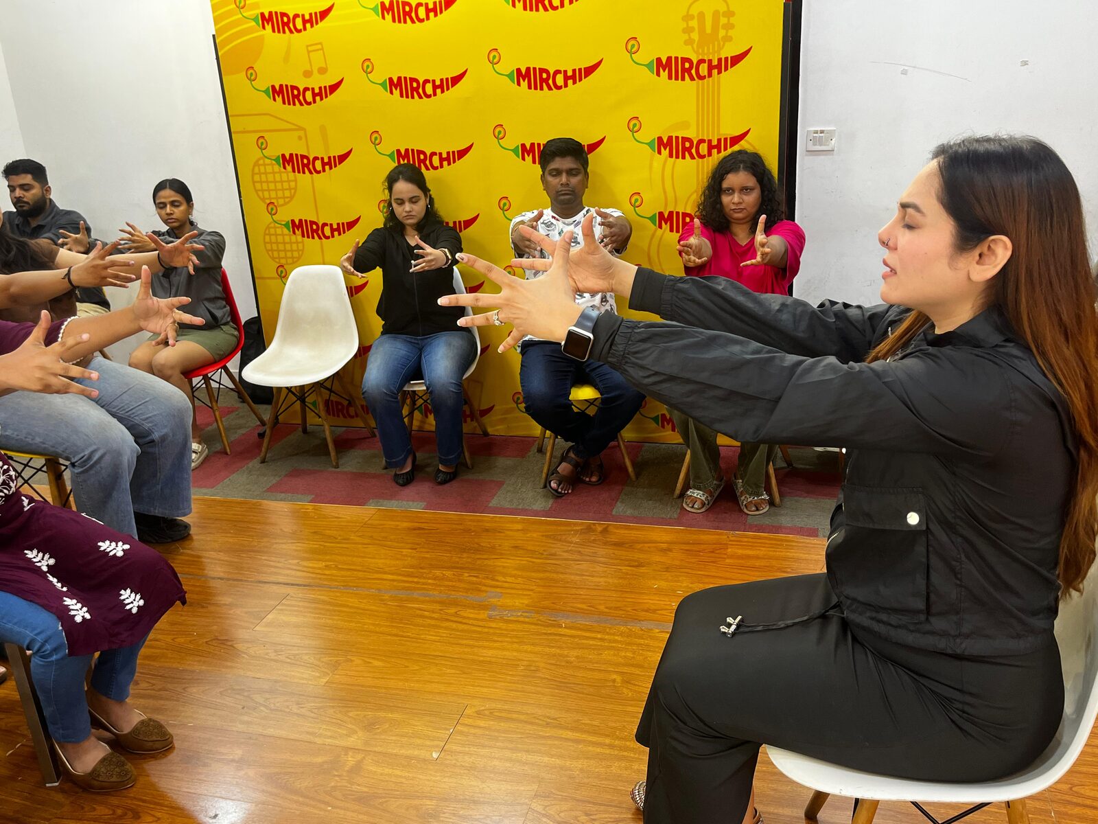 Vibha Jain sits facing a group of seated participants during a workshop, guiding them through an exercise with outstretched arms.